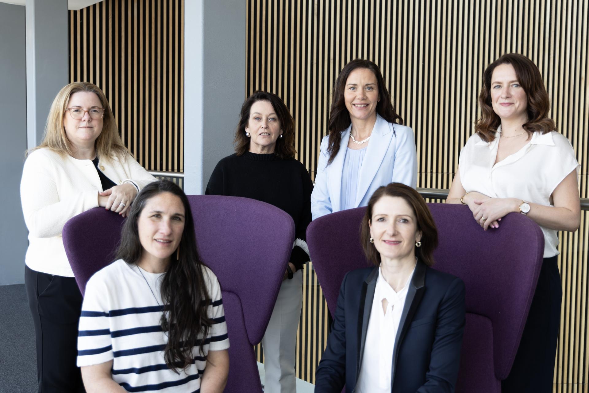 Group portrait of six people in a modern office space. Four people stand behind two high-backed purple chairs while two people sit in front. The background features floor-to-ceiling vertical wooden slats and large windows providing natural light.